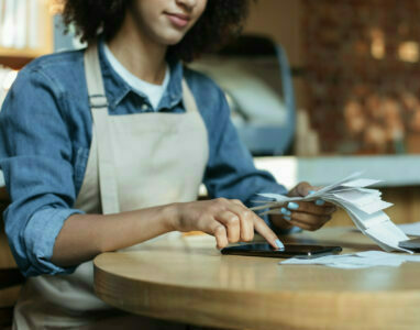 Small business management and accounting. Busy african american young lady owner in apron works with papers, counts costs and profits in interior of modern cafe, cropped, close up, copy space