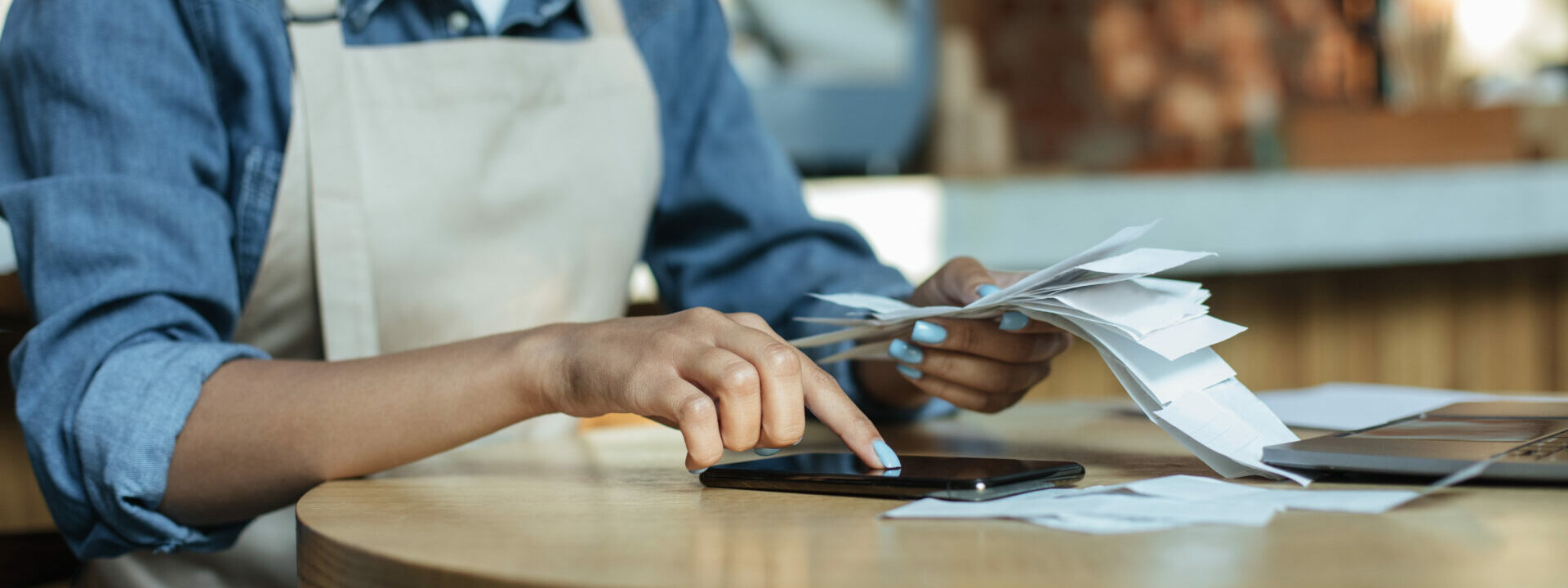 Small business management and accounting. Busy african american young lady owner in apron works with papers, counts costs and profits in interior of modern cafe, cropped, close up, copy space