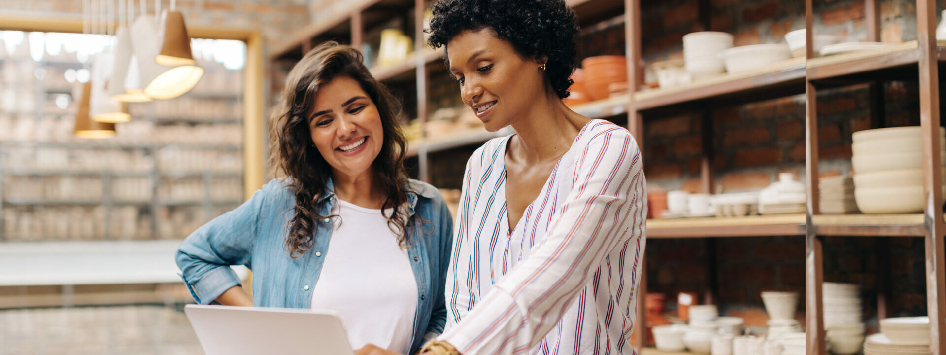 Successful female ceramists using a laptop while working together. Two female entrepreneurs managing online orders in their store. Happy young businesswomen running a creative small business together.