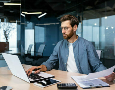 A serious young man accountant, financier, analyst, auditor sits in the office at the table. He holds documents and a pen in his hands, checks accounts, finances, types on a laptop.